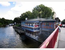 Backsteingebäude mit blauen Holzakzenten an einem Kanal, mit einer Terrasse geschmückt mit Blumenkästen und Lampen. Im Hintergrund sind dichte grüne Bäume, ein vorbeifahrendes Boot und eine belebte Straße mit Passanten und Fahrzeugen zu sehen.