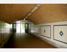 Ein gefliester Fußgängertunnel mit gewölbter Decke aus gelben Klinkersteinen und Wandverkleidungen aus weißen Fliesen mit blauen Mustern. Am Ende des Tunnels befinden sich Metalltüren, durch die Tageslicht und ein Blick ins Freie mit Pflanzen sichtbar sind.