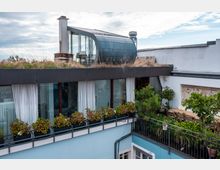 Modernes Gebäude mit Flachdach, begrüntem Dachbereich und einer auffälligen, geschwungenen, verglasten Struktur. Das Obergeschoss verfügt über bodentiefe Fenster mit weißen Vorhängen und einem Balkon mit Pflanzen sowie einem Sitzbereich mit Holztisch und Stühlen.