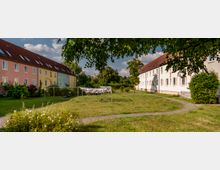 Zwei parallel verlaufende Reihen von pastellfarbenen Wohnhäusern mit Satteldächern umgeben eine grüne Wiese mit einem Wäscheplatz und geschwungenen Gehwegen. Im Hintergrund sind Bäume und eine Hecke zu sehen, die den Bereich abgrenzen.
