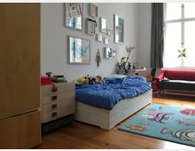 Helles Kinderzimmer mit Holzboden, einem weißen Bett mit blauer Bettwäsche und Kuscheltieren. An der Wand hängen mehrere Bilder, daneben ein Schreibtisch mit Schreibutensilien, ein rotes Sofa sowie ein großer Teppich mit bunten Fischmotiven.