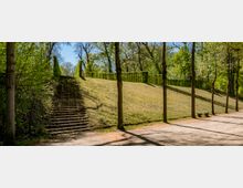 Tree-lined park hillside with broad stone staircases on both sides leading up to a plateau bordered by neatly trimmed hedges. A wide gravel path runs at the base of the slope, with a simple bench and tall trees casting long shadows.
