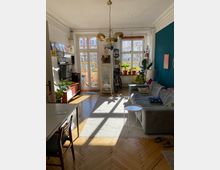 Ein helles Wohnzimmer mit Holzboden und Stuckdecke. Großzügige Fenster und Balkontüren lassen viel Tageslicht herein, ein graues Sofa, ein Esstisch, Zimmerpflanzen und Bücherregale prägen den Raum, während eine goldfarbene Hängelampe in der Mitte hängt.