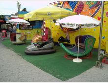 Kleiner Jahrmarktbereich mit Karussellfahrzeugen, darunter ein silberner Autoscooter, eine grüne Wippe und eine rote Sitzgondel, auf grünem Kunstrasen. Im Hintergrund eine gelb gestaltete Wand mit bunten Mustern und ein Verkaufsstand mit Sonnenschirmen.