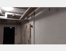 Ein Kellerflur mit sichtbaren weißen Rohren an der Decke, links ein dunkler Durchgang, rechts eine verschlossene Metalltür mit einem gelben Warnschild und darüber eine rote Leuchte. Die Wände sind weiß verputzt und leicht fleckig.