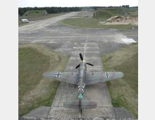 Ein historisches Militärflugzeug auf einem Beton-Flugfeld, umgeben von einer grasbewachsenen Landschaft mit vereinzelten Gebäuden und Hügeln im Hintergrund. Das Flugzeug hat ein Tarnmuster und deutsche Hoheitszeichen, der Blick zeigt es von hinten.