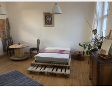 Minimalistisch eingerichtetes Zimmer mit Holzdielenboden. Im Vordergrund ein Bett auf Paletten, im Hintergrund ein runder Tisch aus einer hölzernen Kabeltrommel, zwei Stühle, eine Zimmerpflanze und ein Schrank; an der Wand ein Bild sowie eine teilweise freigelegte Ziegelmauer.