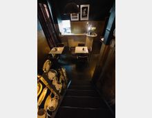 Ein kleines Café mit dunkler Inneneinrichtung, schwarzen Wänden und goldenen Akzenten. Im Raum stehen quadratische Tische mit Stühlen, und an der Treppe im Vordergrund ist ein Regal mit dekorativen Tellern angebracht.