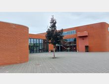 Backsteinbau mit großen Glasfronten, moderner Architektur und abgerundeten Wandelementen. Im Vordergrund ein gepflasterter Platz mit einem kleinen Baum in der Mitte.