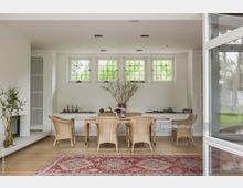 Heller Essbereich mit einem Holztisch und geflochtenen Stühlen, großen weißen Fenstern mit Sprossen, einem roten Teppich und Dekorationen wie Vasen und Pflanzen. Im Hintergrund ist eine weiße Wand mit eingebautem Regal und einer Wandheizung zu sehen.