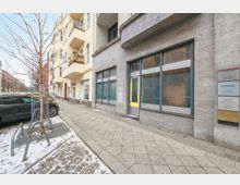 Straßenszene mit breitem Gehweg vor einem grauen Gebäude mit großer Schaufensterfront und Eingangstür sowie einem Schild an der Fassade. Entlang der Straße stehen parkende Autos und ein kahler Straßenbaum, am Boden liegen stellenweise Schnee und Pflastersteine.