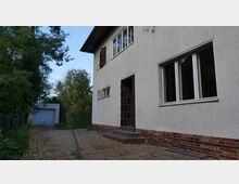 Zweistöckiges Wohnhaus mit hellem Putz und Klinkersockel, umgeben von Bäumen. Der Eingangsbereich mit einer kleinen Treppe ist auf der rechten Seite sichtbar, im Hintergrund eine Garage mit weißem Tor.