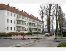 Mehrfamilienhaus mit weißen Fassaden und roten Ziegeldächern, daneben eine mit Bäumen gesäumte Straße. Im Vordergrund befinden sich Baustellenabsperrungen und Verkehrszeichen an einer teilweise gesperrten Straße.