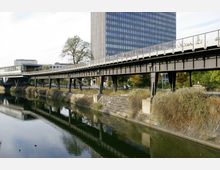 Brücke über einen Kanal mit Stahlkonstruktion, die von Pfeilern getragen wird, im Vordergrund Wasser mit Uferbefestigungen aus Stein. Im Hintergrund ein modernes Hochhaus mit Glasfassade und Bäume.