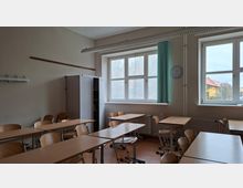 Leeres Klassenzimmer mit mehreren Holztischen und Stühlen, einem hohen Schrank und einer Garderobenleiste an der Wand. Große Fenster lassen Tageslicht herein; daneben hängen eine Uhr und ein grüner Vorhang, unter den Fenstern befinden sich Heizkörper.