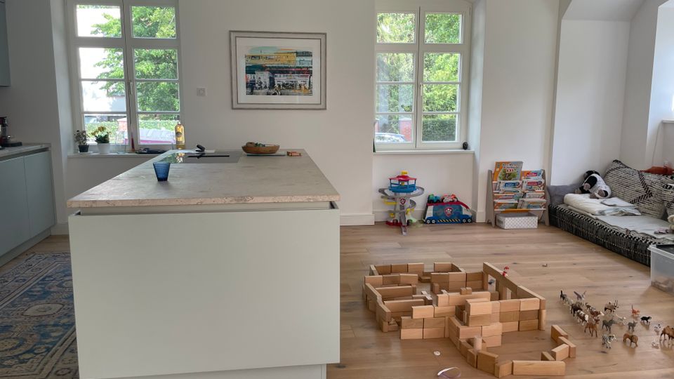 Heller Wohn- und Küchenbereich mit großer Kücheninsel und zwei hohen Sprossenfenstern, durch die Bäume im Außenbereich sichtbar sind. Auf dem Holzboden liegt Spielzeug mit Holzbausteinen, daneben ein niedriges Sofa in einer Nische sowie ein Bücher- und Spielbereich unter dem rechten Fenster.