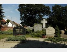 Ein Friedhof mit mehreren Grabsteinen, darunter ein schwarzer Stein mit goldener Schrift und ein großes steinernes Kreuz im Hintergrund. Im Umfeld stehen Bäume, Sträucher und niedrige Pflanzen, mit einigen Wohnhäusern und einer Backsteinmauer im Hintergrund.