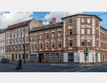 Straßenszene mit historischen Wohn- und Geschäftsgebäuden aus rotem und weißem Ziegel, verziert mit Stuckelementen. Vor den Gebäuden verlaufen Straßenbahnschienen, Gehwege und eine Frau mit Einkaufswagen überquert die Straße.