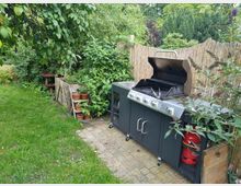 Ein Garten mit einer gepflasterten Ecke, auf der ein Gasgrill mit geöffnetem Deckel steht. Im Hintergrund sind Bambuszäune, grüne Büsche und ein Kompostbereich mit einigen Tontöpfen zu sehen.