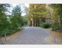 Gepflasterter Weg in einem Park mit Bäumen und Büschen, der zu einem Gebäude mit großer Holztür und Säulen führt; rechts stehen eine Laterne und ein angelehntes Fahrrad.