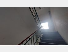 Blick auf ein schlichtes Treppenhaus mit dunklen Stufen, einem Metallgeländer mit rotem Handlauf und einer großen Fensteröffnung, die Tageslicht hereinlässt. Die Wände sind weiß und wirken unrenoviert.