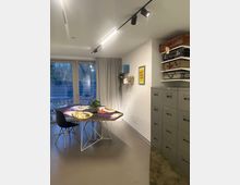 Moderner Arbeits- oder Besprechungsraum mit einem sechseckigen Tisch, darauf eine Pflanze und dekorative Objekte. Im Hintergrund eine große verglaste Tür mit Vorhängen, rechts eine Wand mit grauen Metallschränken und gestapelten Vintage-Koffern sowie einer bunten Wanddekoration.