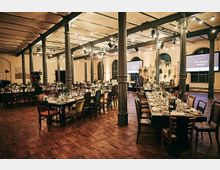 Ein großer, festlich dekorierter Veranstaltungssaal mit Holzboden und gusseisernen Säulen, in dem mehrere lange und runde Tische mit Geschirr, Gläsern und Kerzen gedeckt sind; im Hintergrund große Fenster und eine Bühne mit Bildschirmen.