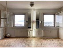 Minimalistischer Innenraum mit weiß gestrichenen Wänden, zwei geöffneten Fenstern und Holzboden. An der Wand befinden sich Heizkörper, schmale Spiegel und kleine Lautsprecher; eine Discokugel hängt zentral von der Decke.