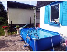 Ein Hinterhof mit einer Terrasse aus Steinplatten, einem blauen rechteckigen Aufstellpool mit Leiter, und einem Haus mit weißer Fassade und blau gestrichenen Fensterläden. Im Hintergrund stehen ein Garten mit Bäumen, Blumen in Pflanzgefäßen und eine Hängematte.