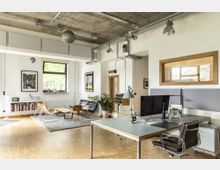 Modernes Büro mit offener Gestaltung: Ein großer Raum mit sichtbarer Betondecke und offenen Lüftungsrohren. Links im Raum stehen eine Couch, ein Teppich und eine Sitzgruppe, während sich rechts ein Schreibtisch mit Monitor und Bürostuhl befindet. Bücherregale, Pflanzen und große Fenster verleihen dem Raum eine helle und einladende Atmosphäre.