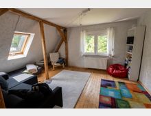 Helles Dachzimmer mit schrägen Wänden, sichtbaren Holzbalken und Holzfußboden, ausgestattet mit Sofa, Sessel und Teppichen. Ein Dachfenster und ein großes Fenster mit Vorhängen lassen Tageslicht herein; daneben stehen ein Regal und ein roter Sitzsack.