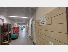 Ein heller Krankenhausflur mit gefliesten Wänden und grauem Bodenbelag, ausgestattet mit Türen zu verschiedenen Räumen. Ein Schild mit der Aufschrift „SYNLAB Blutdepot“ und einem Pfeil weist auf eine Richtung hin; links stehen Transportwagen und Kisten.