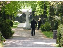 Ein Friedhof mit einem gepflasterten Weg, gesäumt von grünen Bäumen und Sträuchern. Im Hintergrund steht ein großes steinernes Kreuz, und mehrere Personen in schwarzer Kleidung gehen entlang des Weges.