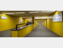 U-Bahn-Gang mit gelb gefliesten Wänden und Deckenbeleuchtung; am linken Rand ein Schild mit der Aufschrift "U7" und Pfeil, sowie ein Handlauf, der zu einem Treppenaufgang führt. Rechts hängen Schilder mit Ausgangs- und Zielinformationen.