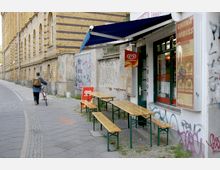 Eine Seitenstraße mit einem gelben Backsteingebäude auf der linken Seite und einem kleinen Restaurant mit Schildern „China Imbiss“ auf der rechten Seite. Vor dem Restaurant stehen Holzbänke und Tische unter einer blauen Markise, während die Wände mit Graffiti bedeckt sind.
