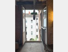 Ein offenes Tor oder Fenster in einem Backsteingebäude mit Blick auf die Fassade eines weißen Wohngebäudes mit mehreren Fenstern. Ein grüner Flaschenzug hängt an der Decke, und auf der rechten Seite befindet sich ein Metallgeländer.