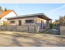 Einstöckiges, modernes Wohnhaus mit Flachdach und beigefarbener Fassade, umgeben von einem Garten. Im Vordergrund ein grauer Lattenzaun mit Tor, und im Hintergrund sind Bäume und angrenzende Gebäude zu sehen.