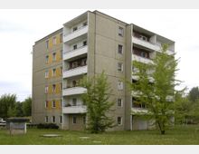 Mehrstöckiges Wohngebäude aus Betonplattenbauweise mit Balkonen auf der Vorderseite, umgeben von einer Grünfläche mit vereinzelten Bäumen. Im Vordergrund steht eine kleine, überdachte Struktur, möglicherweise ein Schaukasten.