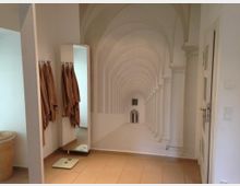 Innenraum eines Badezimmers mit beigefarbenen Fliesen, einem Wandspiegel, an dem Handtücher hängen, und einem Wandbild einer weißen Säulenhalle mit Rundbogenarkaden, das perspektivisch Tiefe erzeugt. Im Vordergrund befindet sich eine Körperwaage.