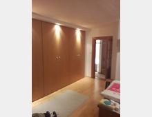 Ein Innenraum mit Holzkleiderschränken an der linken Wand, beleuchtet durch eingebaute Deckenstrahler. Der Raum hat einen hellen Holzfußboden, einen kleinen Teppich in der Mitte und führt zu einer Türöffnung mit einem angrenzenden Flur.