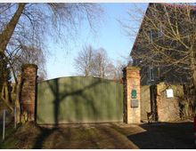 Ein grünes Metalltor ist zwischen zwei gemauerten Backsteinpfosten eingebaut, die zu einer alten Hofeinfahrt gehören. Daneben steht ein graues Gebäude mit roten Dachziegeln, und im Vordergrund sind kahle Bäume sichtbar.