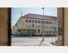 Gebäude mit beiger Fassade und roten Dachziegeln an einer Straßenkreuzung, umgeben von Ampeln und Straßenschildern. Im Erdgeschoss befindet sich ein verglaster Eingangsbereich mit gelben Akzenten.