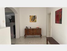 Ein schlichter Innenraum mit weißen Wänden und hellem Marmorboden. Im Vordergrund steht eine antike Holzkommode, über der ein gemaltes Kunstwerk hängt. Rechts befindet sich eine schmale Holzbank, und links führt ein gewölbter Durchgang in einen weiteren Raum.