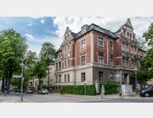 Historisches Gebäude mit roter Klinkerfassade und hellen Steinverzierungen, gelegen an einer Straßenecke mit Bürgersteig, Bäumen und Straßenschildern. Im Vordergrund Fußgänger, parkende Autos und Straßenlaternen.