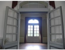 Ein heller Innenraum mit geöffneten Doppeltüren aus Glas und Holzrahmen, die zu einem weiteren Raum mit einem großen Fenster führen. Das Fenster hat einen halbkreisförmigen oberen Teil, dahinter sind rote Vorhänge sichtbar.