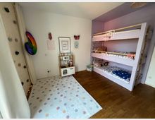 Kinderzimmer mit Holzboden, einem Etagenbett in hellvioletten Farben und einer Kletterwand links mit bunten Klettergriffen. Im Vordergrund liegt ein gepunkteter Teppich, und an der Wand steht eine kleine Spielküche.
