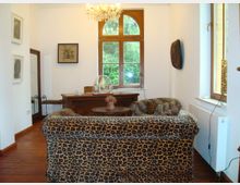 Wohnraum mit Möbeln im Leopardenmuster, einem Holztisch und einem Schreibtisch im Hintergrund. Ein großes, bogenförmiges Fenster mit Blick ins Grüne dominiert die weiße Wand, darüber hängt ein Kronleuchter.