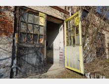 Eingang eines verfallenen Backstein- und Putzgebäudes mit zwei geöffneten, verglasten Holztüren, eine dunkel und eine gelb gestrichen. Vor dem Eingang stehen trockene Sträucher und Äste, der Boden ist teils Beton, teils Gras.