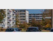 Wohngebiet mit mehrstöckigen Wohnhäusern, darunter ein helles Gebäude mit blauen Balkonen und Blumenkästen. Im Vordergrund sind geparkte Autos, Bäume mit Herbstblättern und rechts ein Straßenschild mit der Aufschrift „Baumschulenweg“ zu sehen.