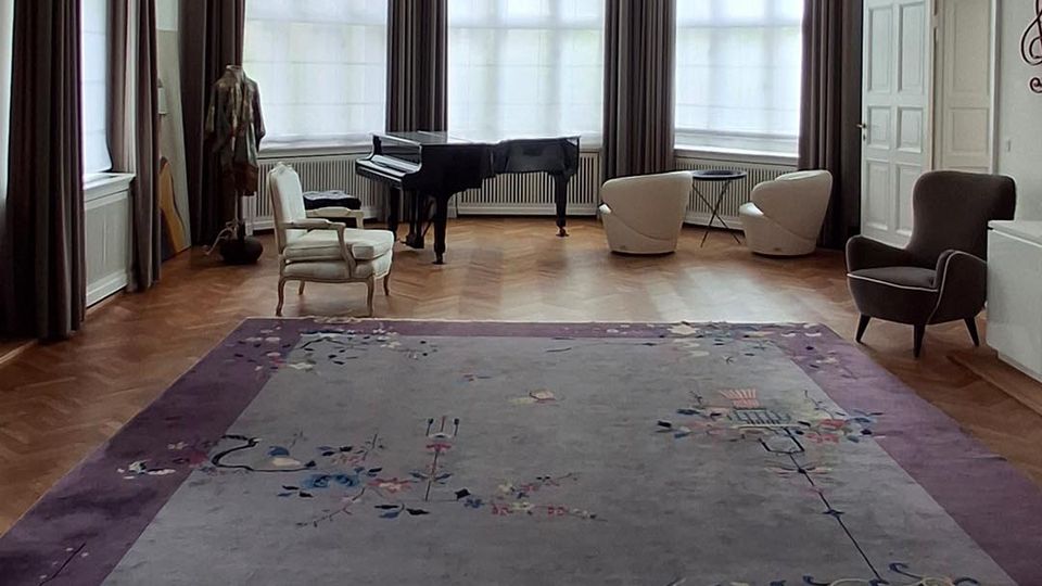 Großer, heller Salon mit hohen Decken, zentralem schmiedeeisernen Kronleuchter und umlaufenden hohen Fenstern mit dunklen Vorhängen. Auf Parkettboden liegt ein großer gemusterter Teppich; an den Wänden stehen ein Flügel und mehrere Sessel.